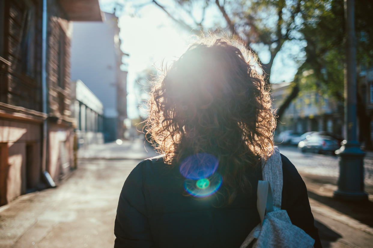 Rear view young woman walking down the street, sun flare towards her. Female with curly hair goes along city sidewalk.