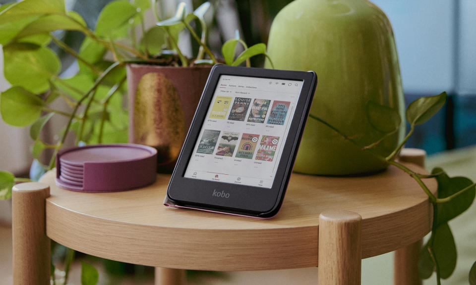 Product lifestyle photography for the Kobo Clara Color e-reader. The device is propped up on a small round wooden table in front of a vase of plants.