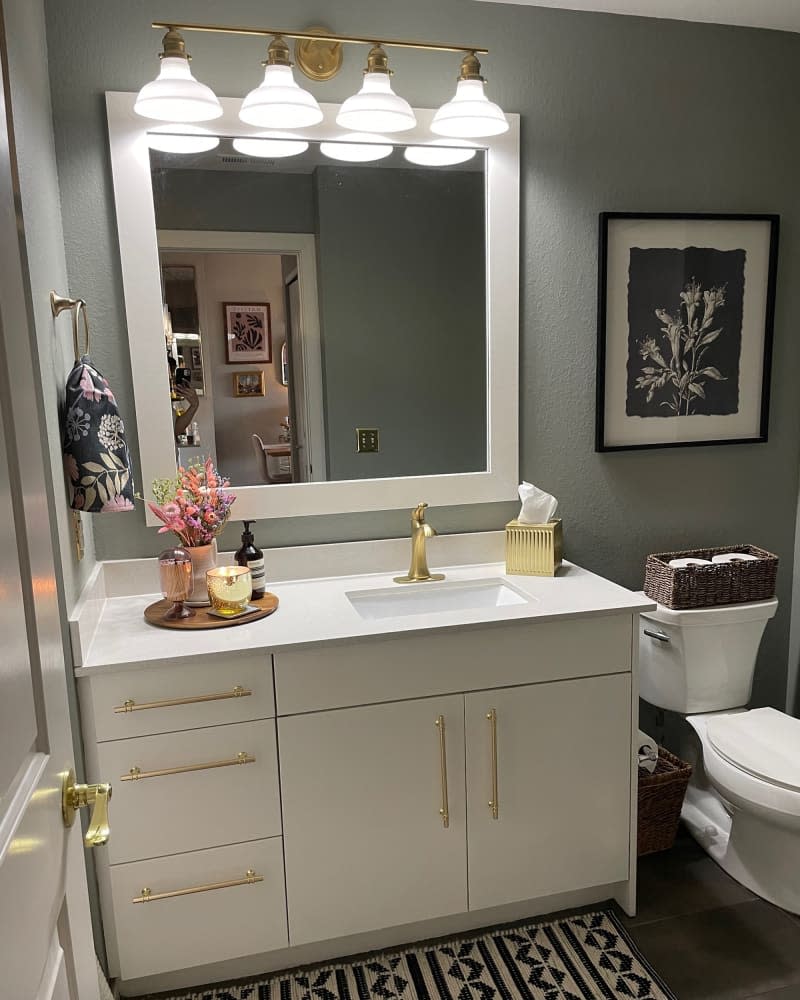 Lights above white framed mirror over white sink with wood tray holding accessories and gold tissue box in sage bathroom with floral towel and art and graphic rug.