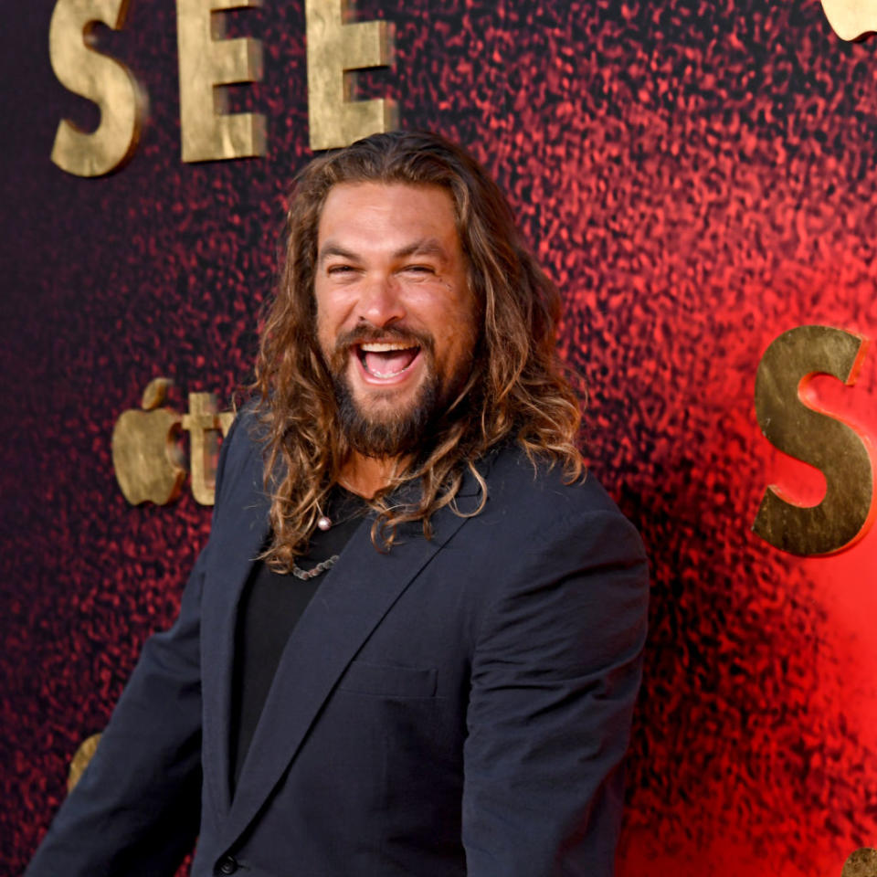 Man in a black suit laughing at a premiere, standing before a promotional backdrop