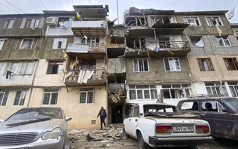 A damaged residential apartment building following shelling is seen in Stepanakert, Nagorno-Karabakh. A man is walking away from the building, which has damage including broken windows, debris-covered balconies and damaged walls.
