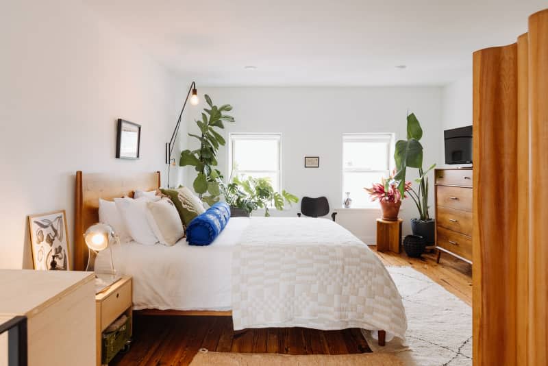 View into bedroom with bed with wood headboard, white linens, blue and green pillows. Lots of plants