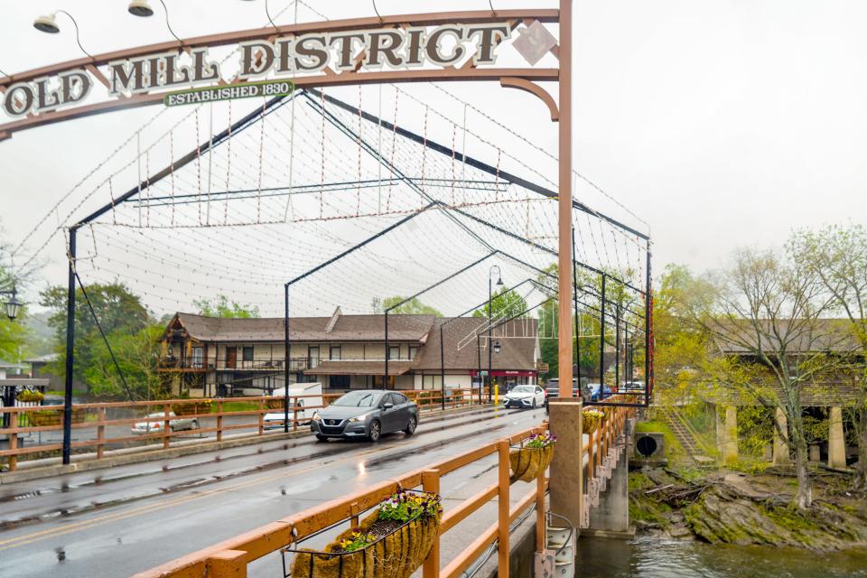 A sign says Old Mill District at the top of a bridge with cars driving under it. Gray skies and trees in the background