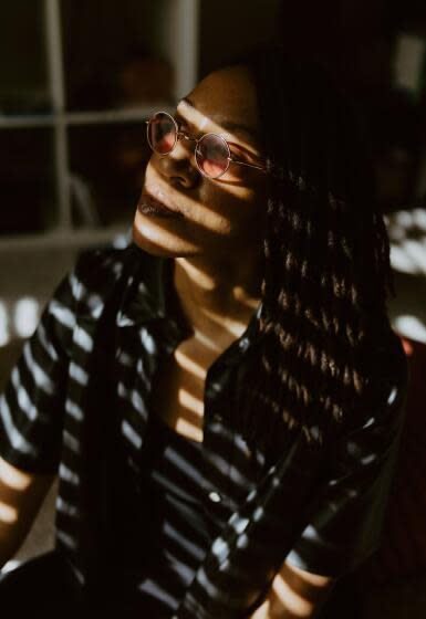 The shadows of a blind cast lines across Vanita Blackburn, a Black woman with shoulder-length locs and round glasses.