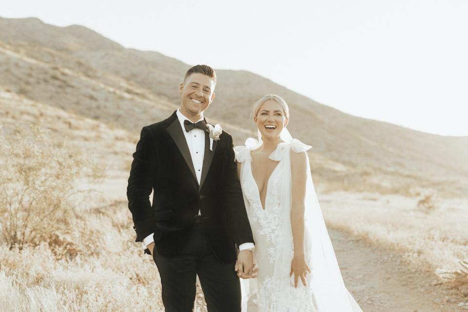 A bride and groom smile and hold hands in front of a hilly backdrop.