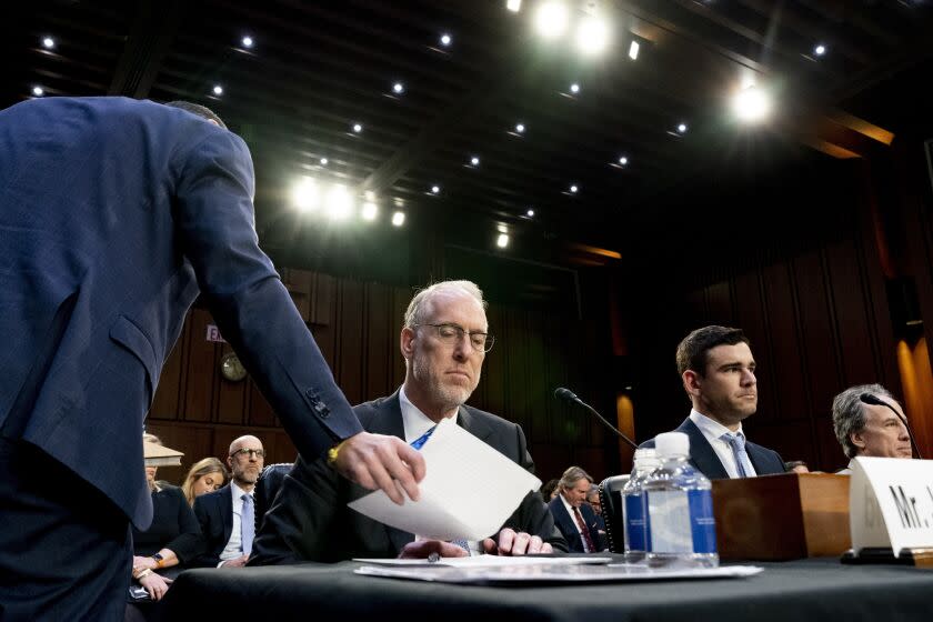 A man in a suit handing a document to another man in a suit seated at a desk inside a courtroom