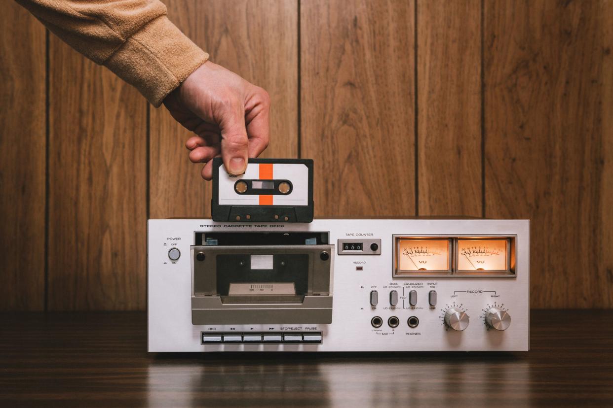 A vintage looking tape player / recorder stereo sits on the counter of a 1970's - 1980's  living room with wood paneling.  A mans hand reaches down to insert a cassette into the machine.  Horizontal image with copy space.