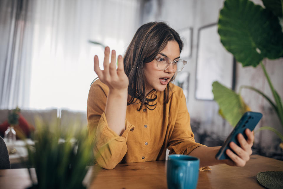 A woman in a mustard top and glasses sits at a table, looking at her phone with a surprised expression, holding her hand up as if questioning something