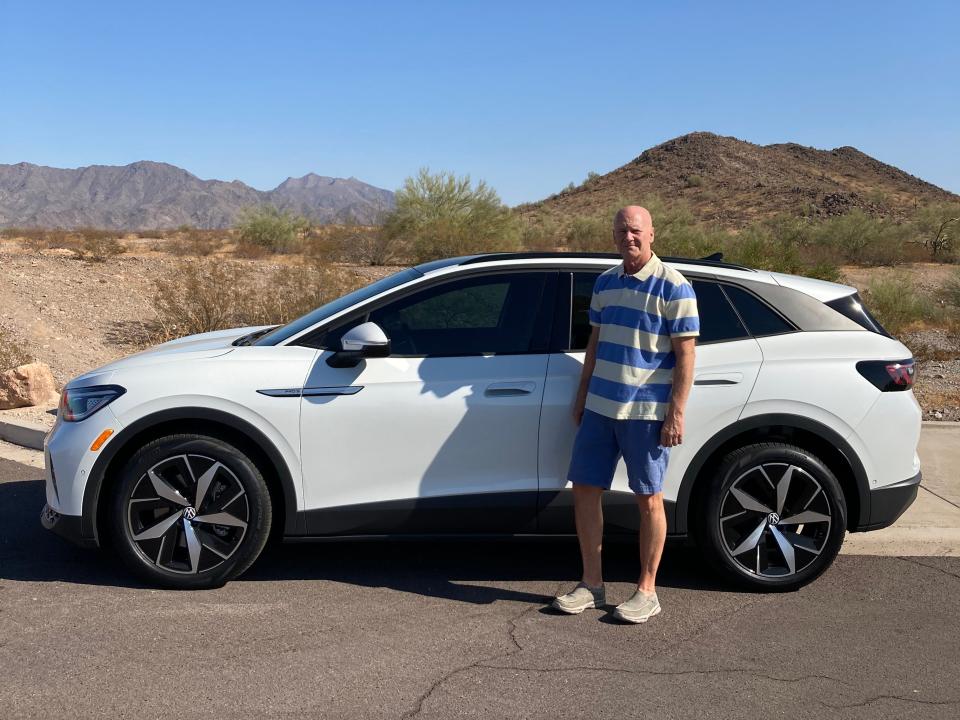 A man stands in front of a white SUV in front of a desert scene.