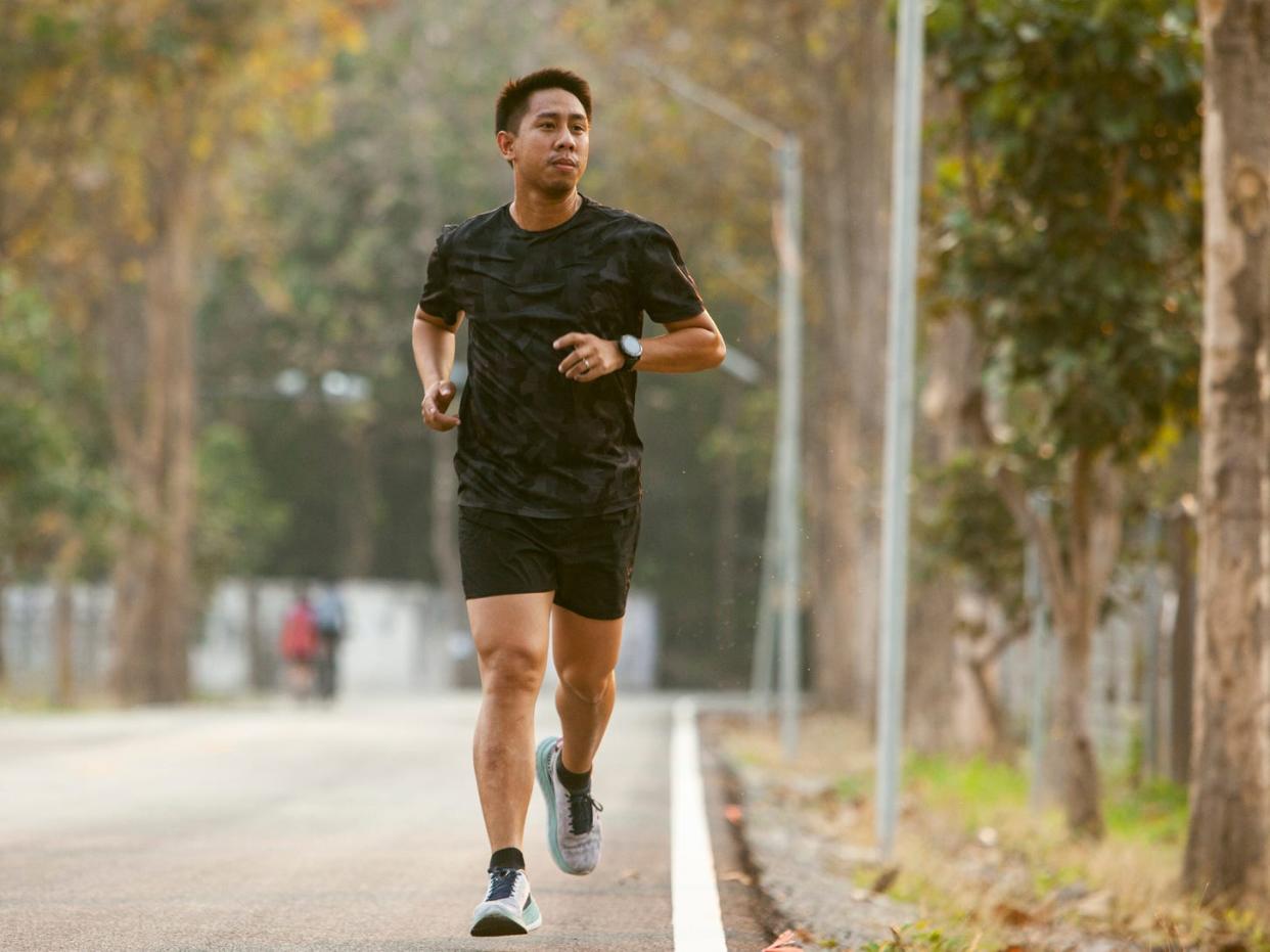 A front view of a man in athletic shorts and a T-shirt jogging down a road surrounded by trees