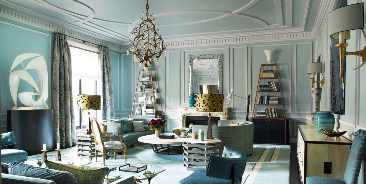 parisian looking space in pale bluish green with high ceilings with moldings and an antler chandelier at center and a blue somewhat geometric rug and modern blue sofa and chairs and demi lune settees and cocktail tables and and a fireplace and mirror and etagere against the back wall