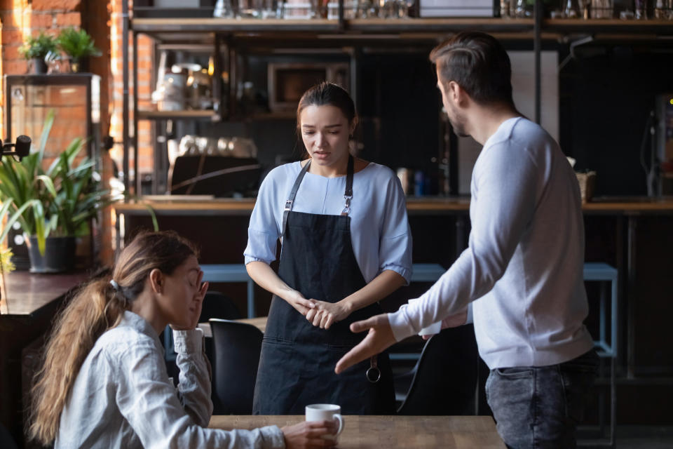 Waitress in apron attending to a seated female customer while a standing male gestures, in a cafe setting