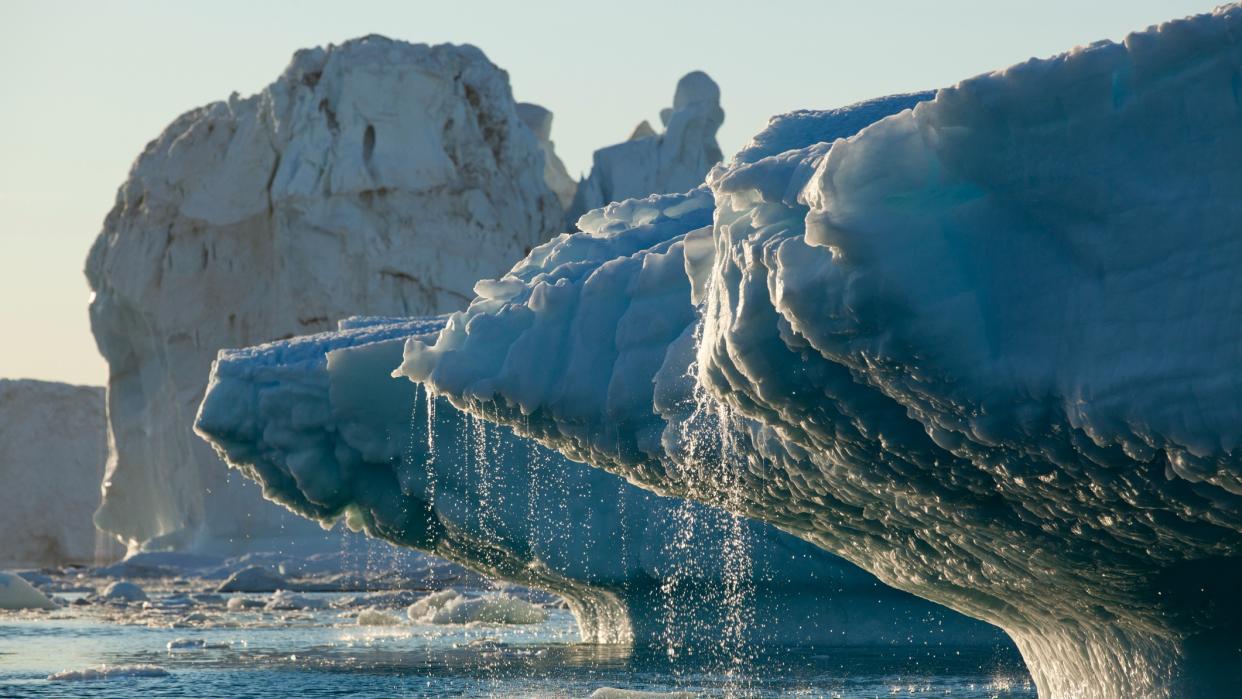  close up of a glacier with melting water running into the water below. other cracked glaciers stand in behind against a pale sky 