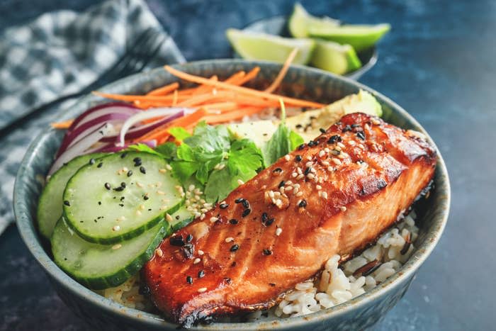 A bowl of rice topped with grilled salmon, sliced cucumbers, red onions, carrots, avocado, and sesame seeds, garnished with fresh herbs and lime wedges on the side