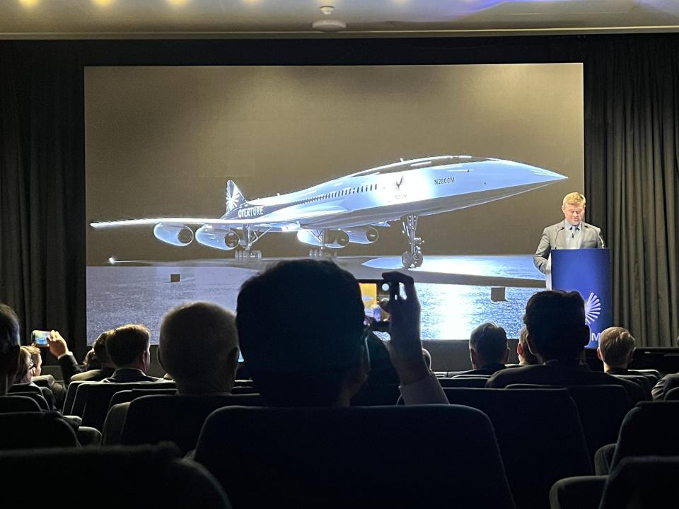 Boom CEO Blake Scholl stands at a podium in a Paris Air Show auditorium, with the overture jet displayed behind him.