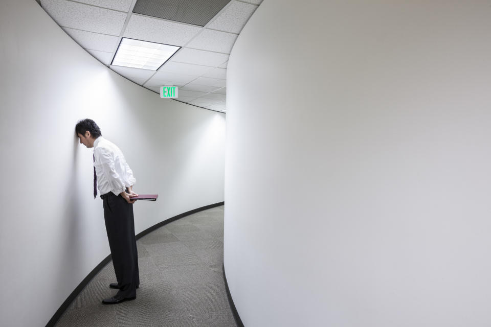 An Asian businessman frustrated and standing with his head against a wall in a hallway.