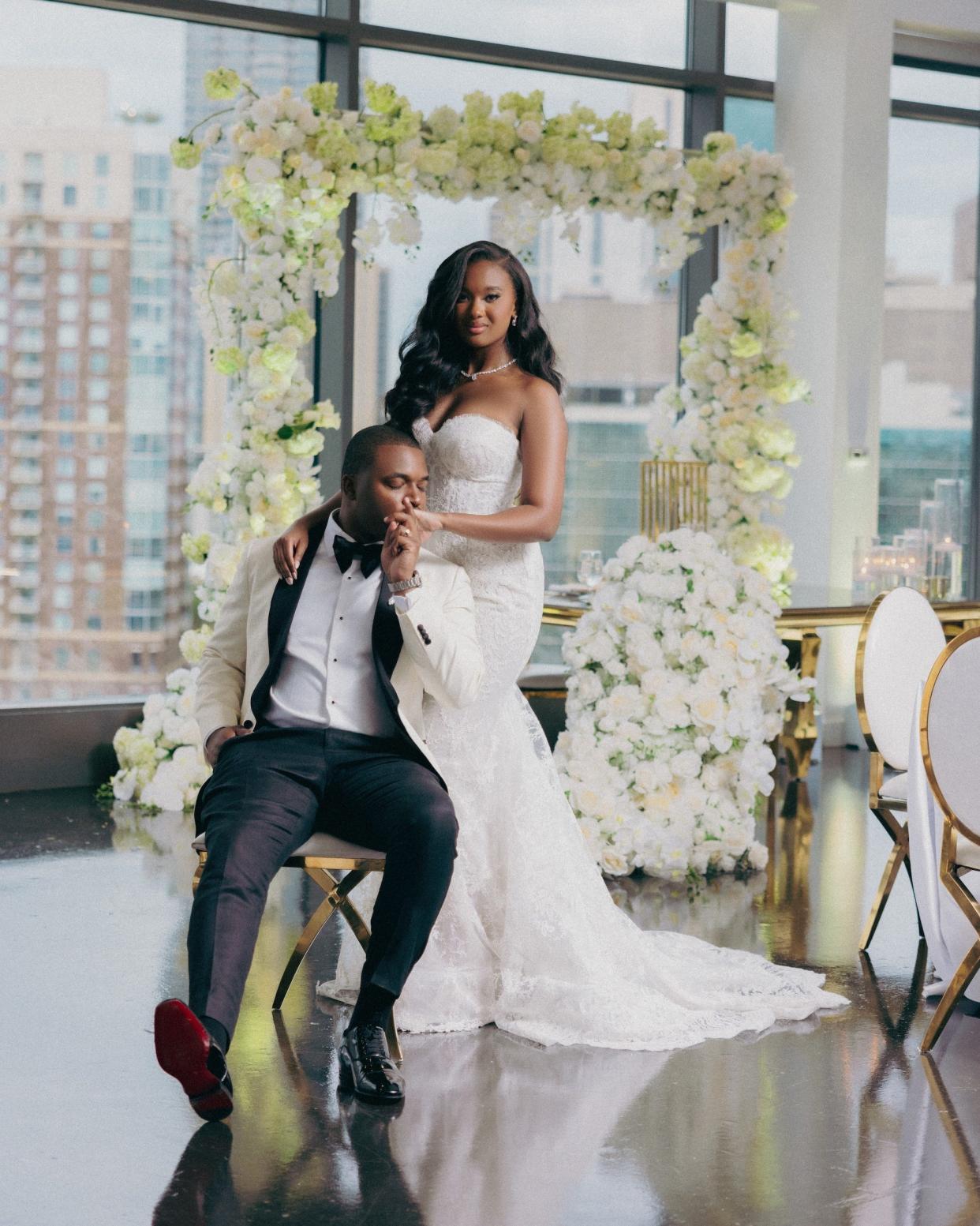 A groom sits in a chair as his bride stands behind him. He kisses her hand.