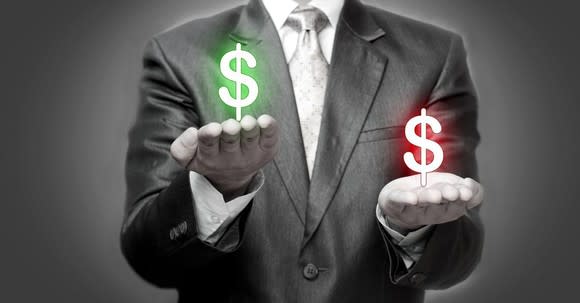 A man in a suit holds a green and red dollar sign in each hand as if weighing them.