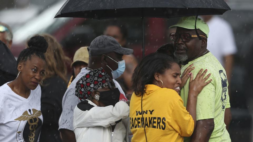 Bystanders gather under an umbrella as rain rolls in after a shooting at a supermarket on Saturday, May 14, 2022, in Buffalo, N.Y.  Officials said the gunman entered the supermarket with a rifle and opened fire. Investigators believe the man may have been livestreaming the shooting and were looking into whether he had posted a manifesto online.