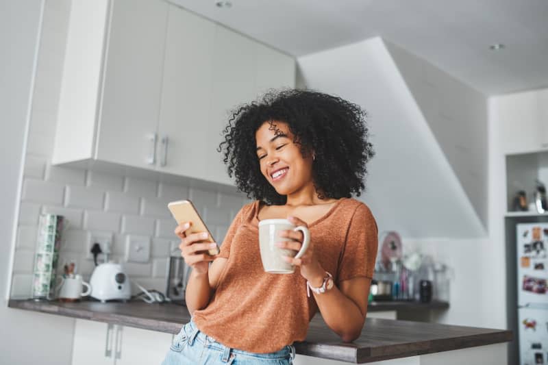 Shot of a young woman using a smartphone and having coffee in the kitchen at home.