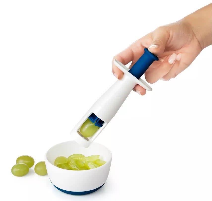 A hand using an OXO Good Grips grape cutter to slice green grapes into a bowl