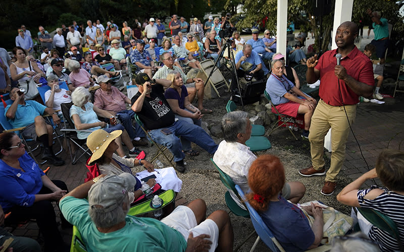 Republican presidential candidate Sen. Tim Scott (R-S.C.) speaks at a barbecue. He is standing to the right of the image, facing a crowd that is seated in a semicircle around him