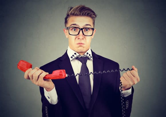 Man in a navy suit, holding the handset of a red landline phone in his hands with a shocked expression on his face.