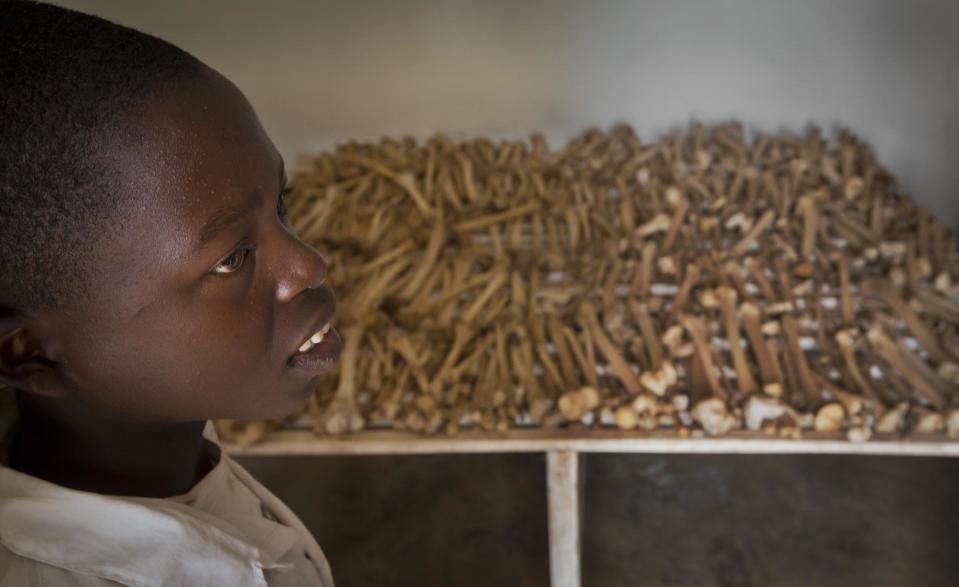 In this photo taken Thursday, March 27, 2014, Nikeyimana Obadia, 12, who lives nearby, stands by rows of human bones that form a memorial to those who died in the redbrick church that was the scene of a massacre during the 1994 genocide, in the village of Nyarubuye, eastern Rwanda. Rwandans gathered in the nearby town of Kirehe Thursday to watch the arrival of a small flame, symbolic fire traveling the country as Rwanda prepares to mark 20 years since ethnic Hutu extremists killed neighbors, friends and family during a three-month rampage of violence aimed at ethnic Tutsis and some moderate Hutus, the death toll of which Rwanda puts at 1,000,050. (AP Photo/Ben Curtis)