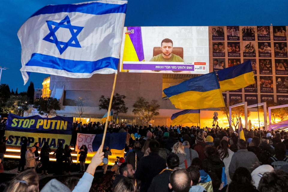 Demonstrators carrying Israeli and Ukrainian flags gather beneath a video screen on which Volodymyr Zelensky appears.