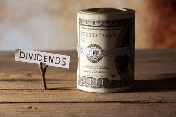 The word dividends written on a piece of paper next to a roll of hundred-dollar bills atop a wooden table.