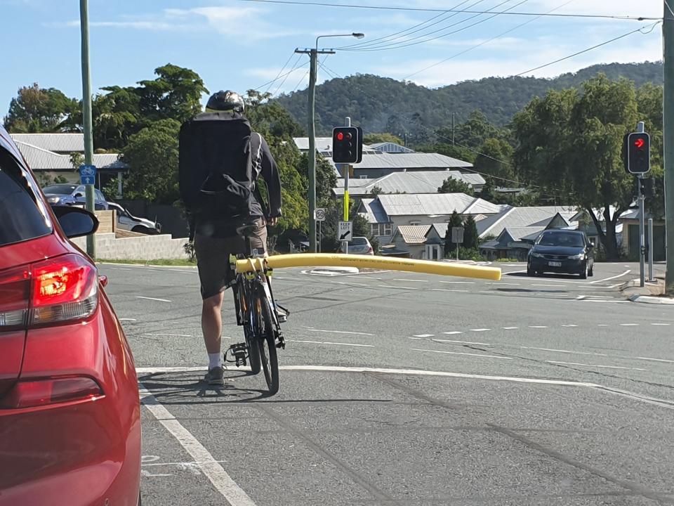 A Brisbane cyclist is stopped at traffic lights with a long yellow pool noodle extending from the right side of his bike. Source: Facebook