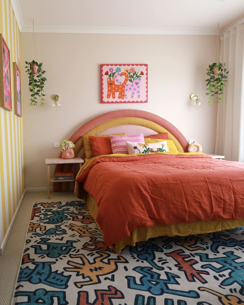 Bedroom with bed with orange and yellow curved fabric headboard, keith haring rug, and yellow and white striped accent wall
