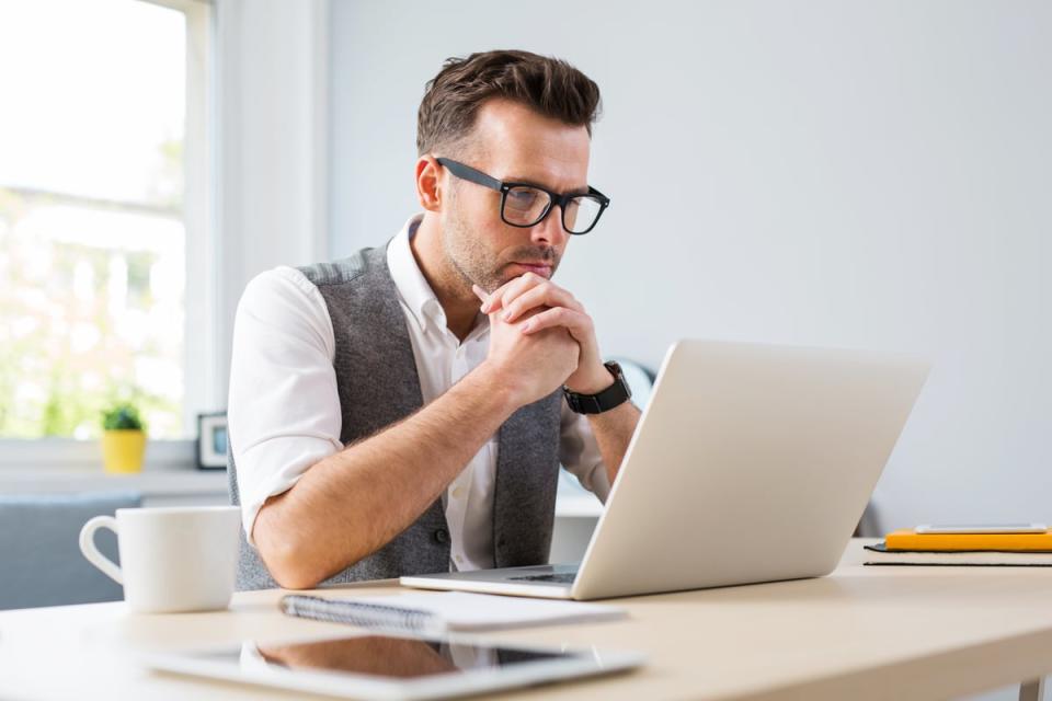 A person clasps their hands while sitting at a table in front of a laptop computer.