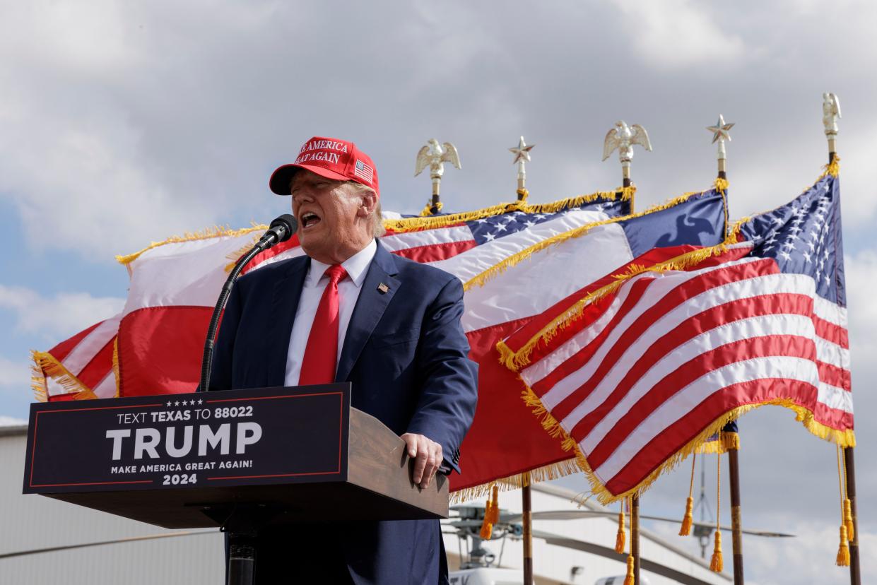 Donald Trump standing behind a podium at a campaign event wearing a MAGA hat.