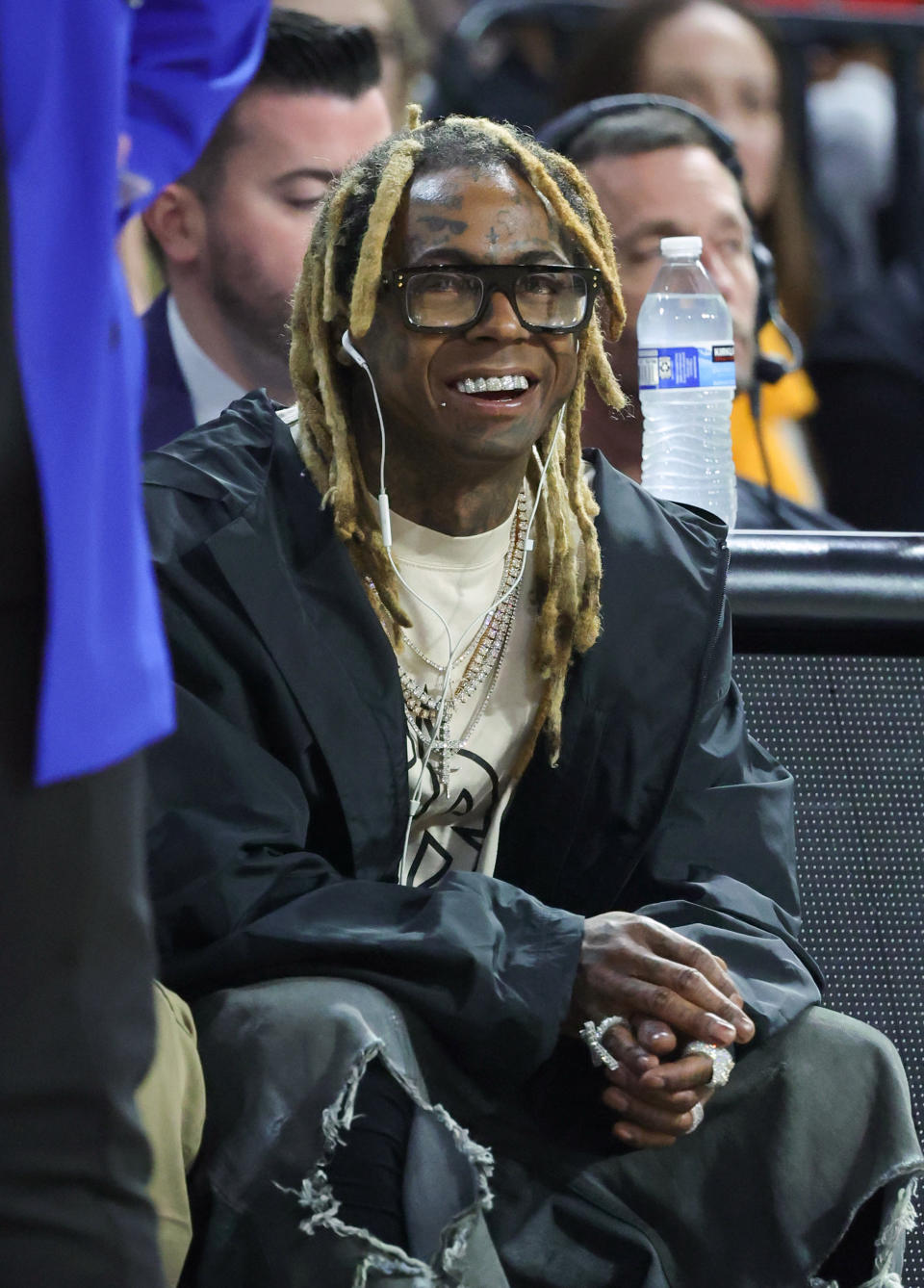 Lil Wayne smiling and sitting courtside at a sports event, wearing a black jacket over a graphic t-shirt, with layered necklaces and multiple rings