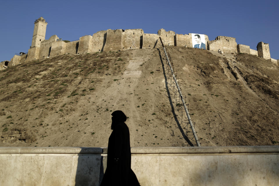 In this Saturday, July 27, 2019 photo, a woman walks in front of the ancient Citadel with a giant picture of Syrian President Bashar Assad, in Aleppo, Syria. Rebels still frequently strike with shelling and mortars into Aleppo, killing civilians nearly three years after the government recaptured the city. Aleppo is a symbol of how President Bashar Assad succeeded in turning the tide in Syria’s long civil war with a series of wins, but it’s equally a symbol of how he’s been unable to secure a final victory. Half of Aleppo remains in ruins, and rebels remain on the doorstep. (AP Photo/Hassan Ammar)