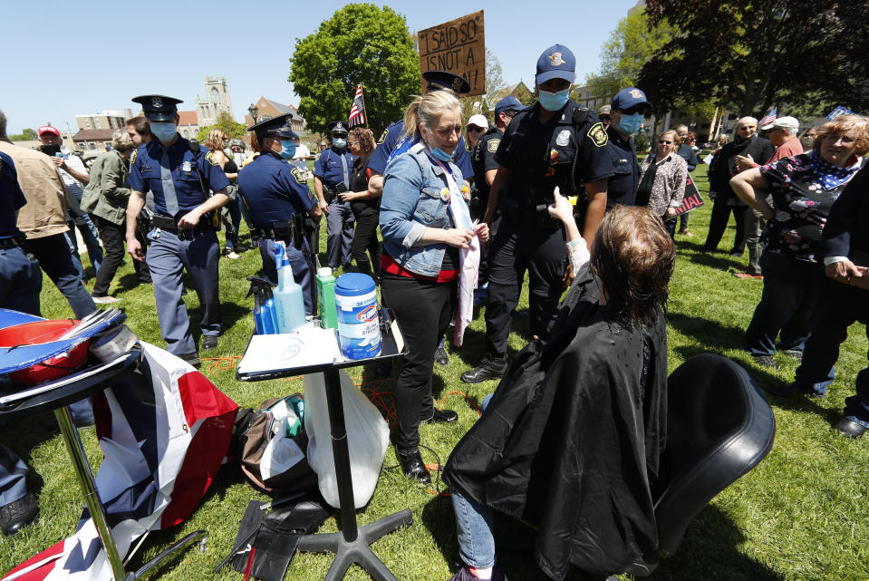 Linda Hicks, seated, points at a Michigan State Police trooper while receiving a free haircut from Teresa Luks, left, at the State Capitol during a rally in Lansing, Mich., Wednesday, May 20, 2020. Barbers and hair stylists are protesting the state's stay-at-home orders, a defiant demonstration that reflects how salons have become a symbol for small businesses that are eager to reopen two months after the COVID-19 pandemic began. (AP Photo/Paul Sancya)