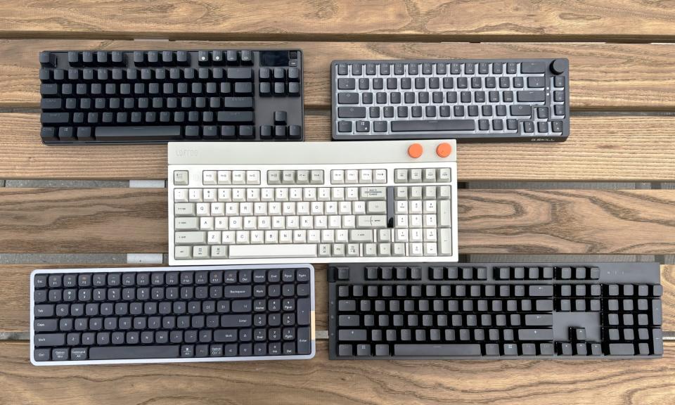 A quintet of mechanical keyboards rest on a brown wooden outdoor table, with one beige model flanked two separate keyboards above it, and two other models below it.