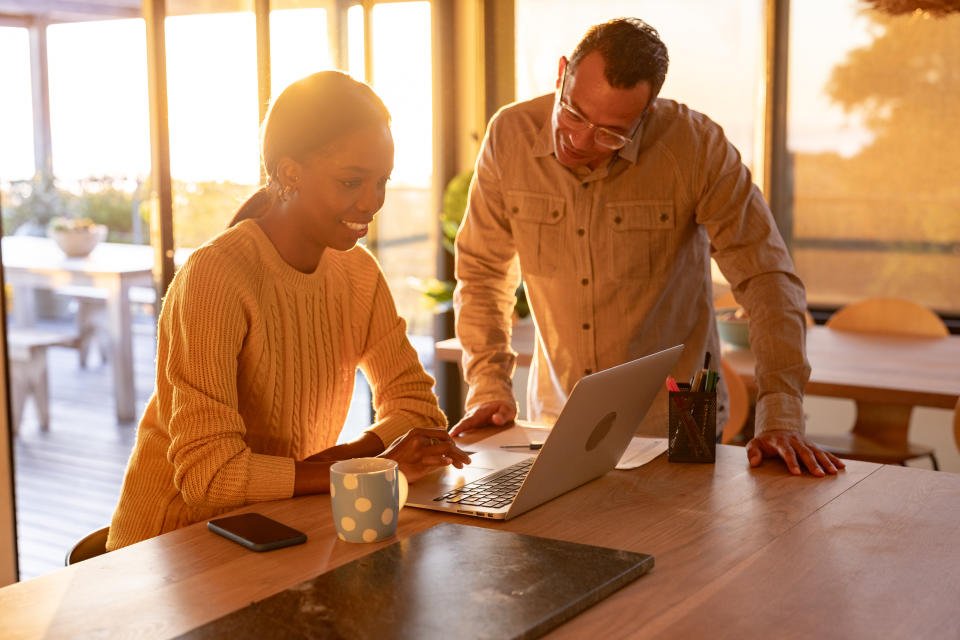 A happy couple sits at their kitchen island looking at a laptop together and going over their self-directed investments while the sun shines in