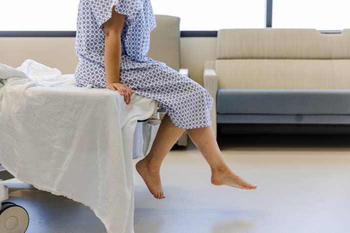 A person in a hospital gown sits on the edge of a hospital bed, with feet dangling above the floor. A waiting area with a sofa is in the background