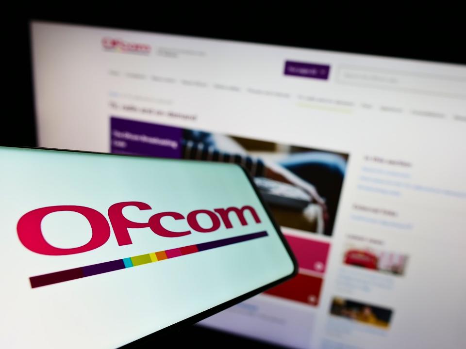 Smartphone with logo of British authority Office of Communications (Ofcom) on screen in front of website. Focus on center-left of phone display.