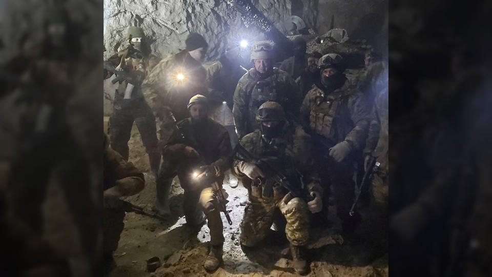 Five members of the Russian mercenary group Wagner pose with Yevgeny Prigozhin underground, lit by strong spotlights. One wears a balaclava, two wear wool hats and two wear helmets, while Prigozhin has a khaki helmet with goggles over it. 