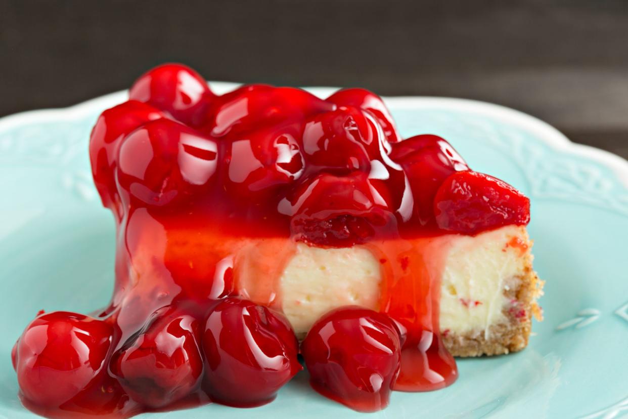 An extreme close up horizontal photograph of a slice of cheesecake topped with a generous portion of glazed cherries.