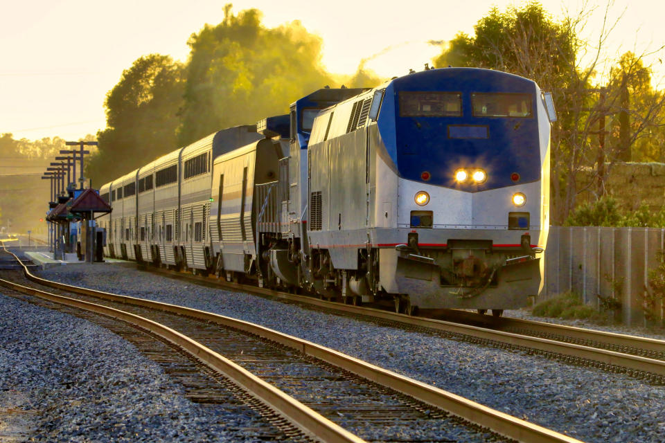 A modern passenger train with multiple cars travels on railroad tracks through a rural area with trees in the background