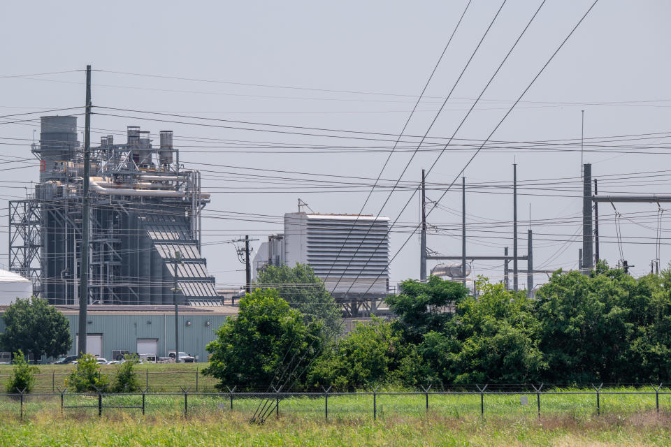 A power facility, surrounded by dozens of power lines, is seen beyond a grassy field with a barbwire fence.