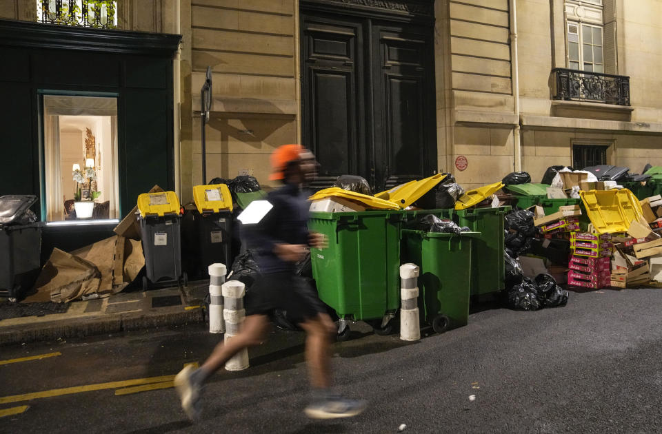 Man jogs past not collected garbage cans in Paris, Monday, March 13, 2023. A contentious bill that would raise the retirement age in France from 62 to 64 got a push forward with the Senate's adoption of the measure amid strikes, protests and uncollected garbage piling higher by the day. (AP Photo/Michel Euler)