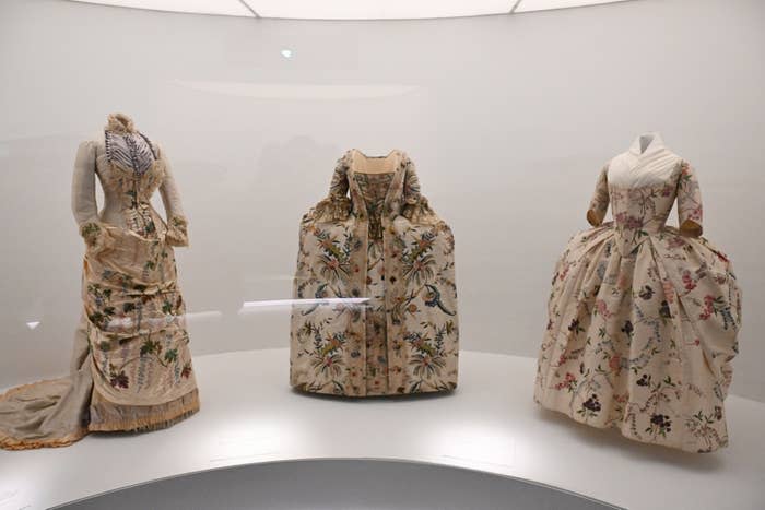 Three historical dresses on display, featuring intricate patterns and full skirts