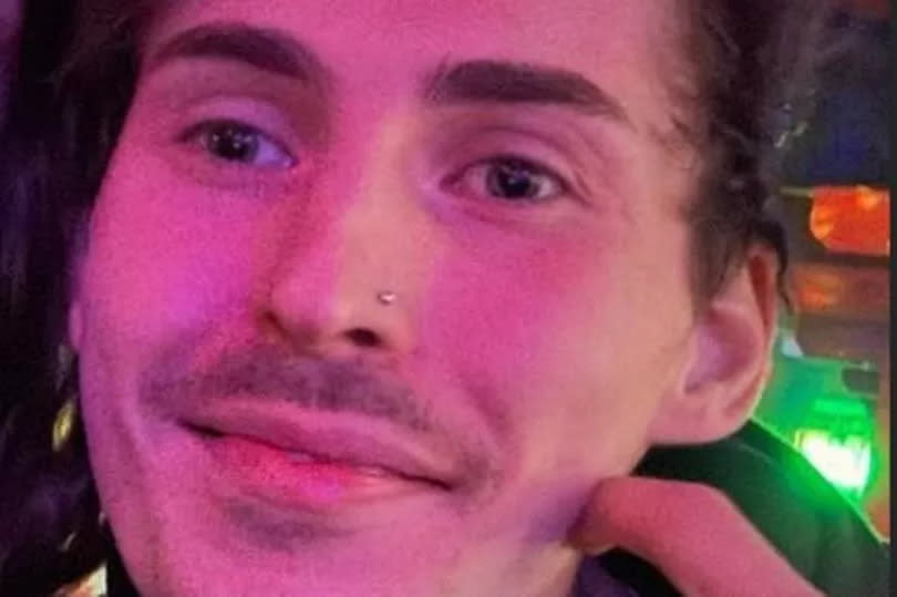 A young man with a slight moustache smiles for the camera as he looks slightly away from the shot. His face is bathed in a purple light as though in a nightclub or bar