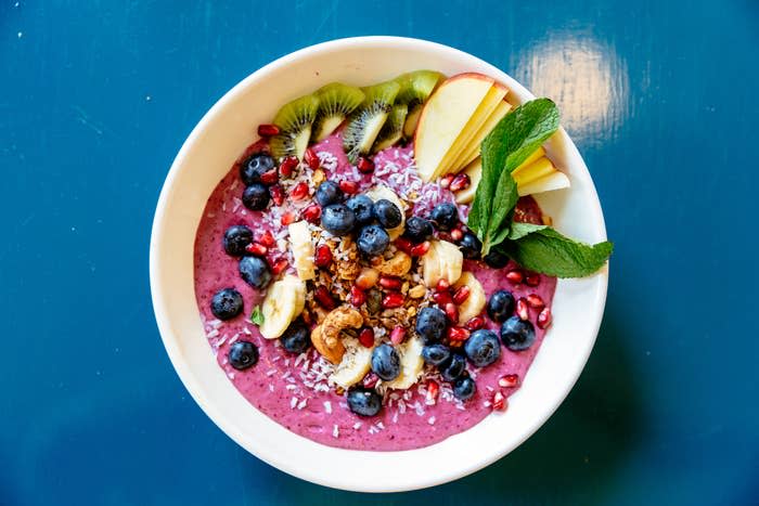 A smoothie bowl topped with blueberries, banana slices, pomegranate seeds, kiwi slices, apple slices, granola, coconut flakes, and mint leaves