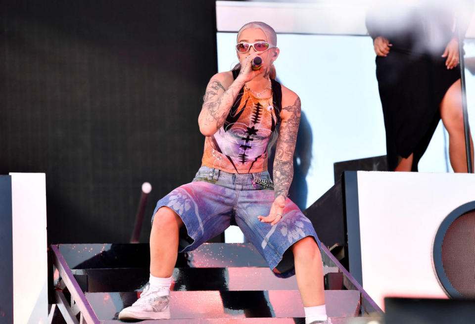Miko in a tank top and sunglasses sings into a microphone onstage with another person partially visible in the background